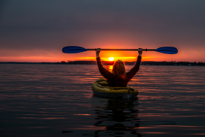 woman in kayak with ore above her head in the sunset