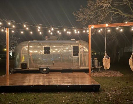 A silver camper illuminated by string lights; next to the camper are two hammocks and an outdoor patio.