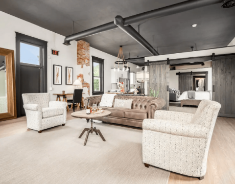 A modern, industrial living room featuring white chairs and a neutral couch; there are high ceilings and natural light.