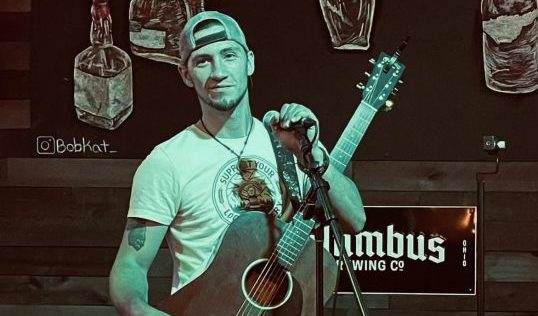 Photo of artist Jarron Sollee posing with a guitar and microphone.