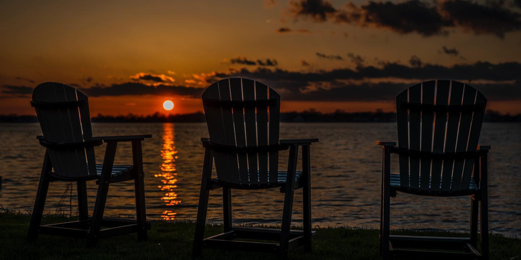 Three chairs sit beside lake as sun is setting.