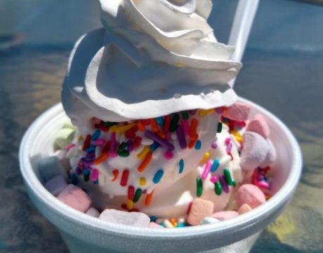 Photo of soft serve ice cream with marshmallows, rainbow sprinkles, and whipped cream on top.